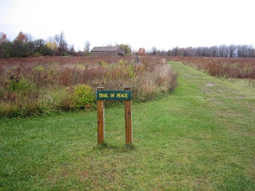 A wooden sign reading "Trail of Peace" stands at the start of a grassy path through a field with autumn foliage.