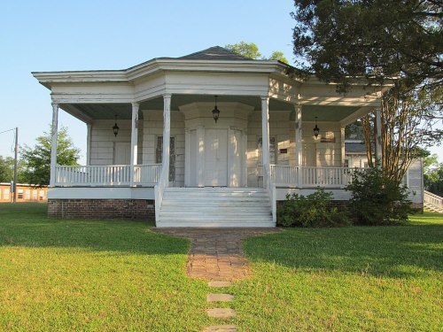 A historic white house with a large porch, surrounded by green grass and trees, under a clear blue sky.