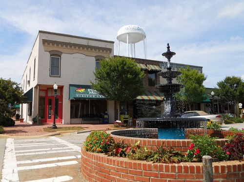 A charming town square featuring a fountain, shops, and a water tower under a blue sky with scattered clouds.