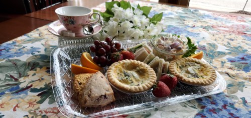 A tray with pastries, fruit, sandwiches, and a cup of tea, surrounded by flowers on a floral tablecloth.