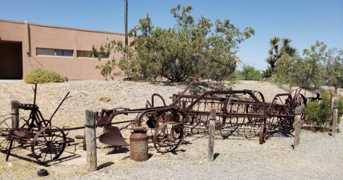Rusty vintage farming equipment displayed outdoors, surrounded by sparse vegetation and a building in the background.