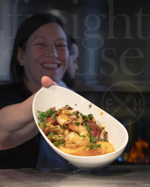 A smiling chef holds a large dish of shrimp and vegetables, showcasing a vibrant meal in a kitchen setting.