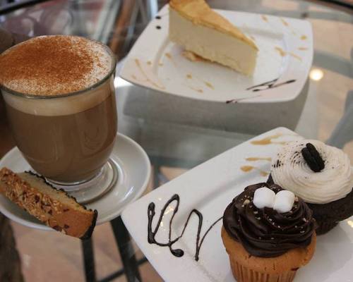 A coffee cup with a biscotti, a slice of cheesecake, and two decorated cupcakes on a glass table.