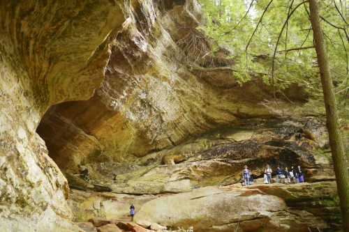 A group of people stands beneath a large rock overhang in a forested area, surrounded by towering trees.