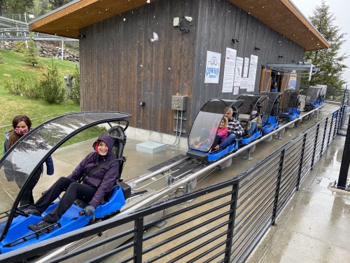 A group of people in blue sleds on a mountain coaster, with a wooden building and snow falling in the background.
