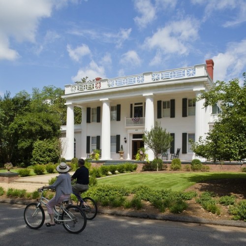 Two people ride bicycles past a large white mansion with columns and a well-maintained lawn under a blue sky.