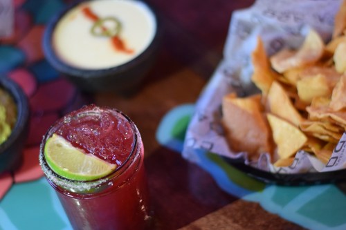 A vibrant cocktail with lime, a bowl of creamy dip, and a plate of crispy tortilla chips on a colorful table.