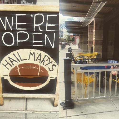 A sign reading "We're Open" outside Hail Mary's, with yellow chairs and tables on a sidewalk patio.