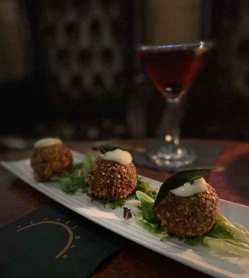 Three crispy appetizers on a white plate, garnished with greens and topped with a dollop of sauce, with a cocktail in the background.