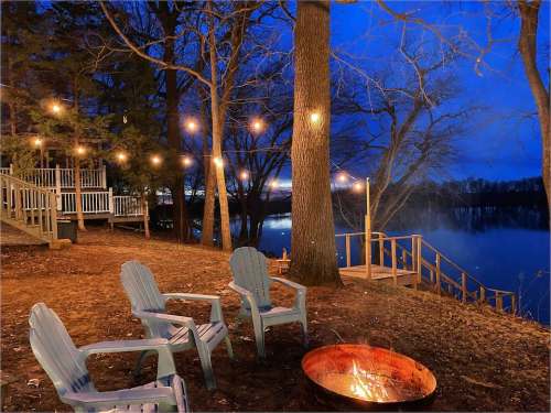 Cozy outdoor scene at night with string lights, a fire pit, and chairs by a calm lake surrounded by trees.