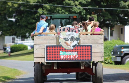 A tractor pulls a wooden wagon with children, featuring a sign for Barstow's and Cabot. Green fields in the background.