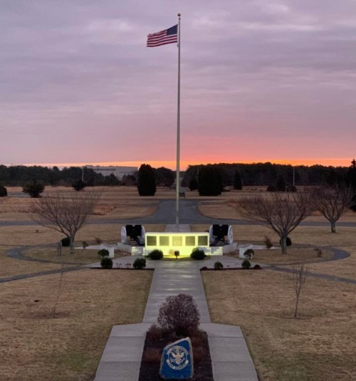 A flagpole with an American flag stands at sunset, surrounded by a memorial and landscaped pathways.