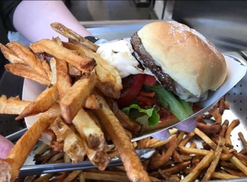A hand holding a burger with lettuce, tomato, and mayonnaise, alongside a serving of crispy French fries.