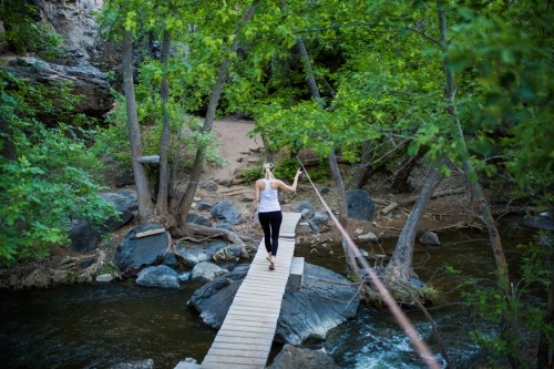 A person walks on a wooden bridge over a stream, surrounded by lush greenery and rocky terrain.