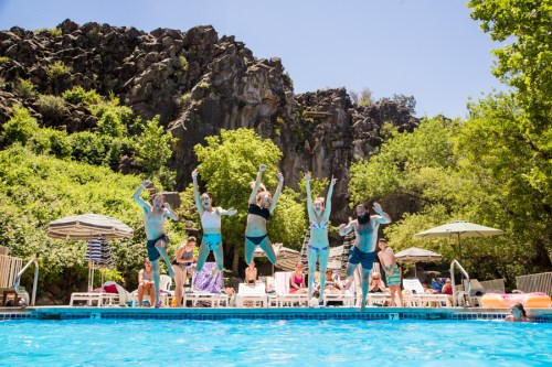 A group of people jumps into a pool, surrounded by greenery and rocky cliffs, enjoying a sunny day.