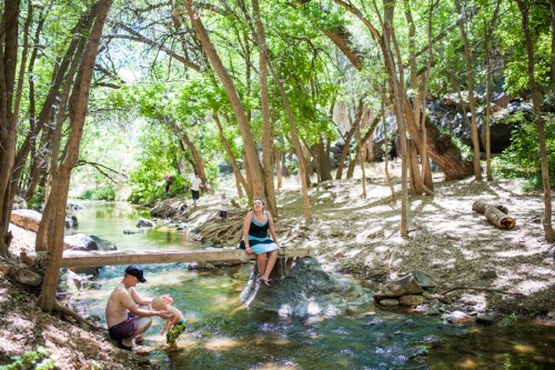 A woman sits on a rock by a stream, while a man wades in the water, surrounded by lush green trees.