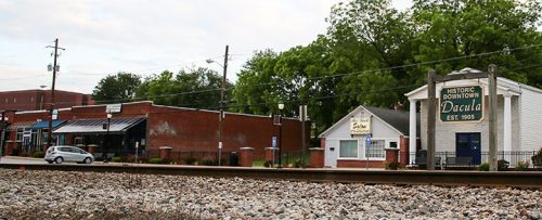 A view of a small town with shops and buildings along a railway track, featuring trees and a cloudy sky.