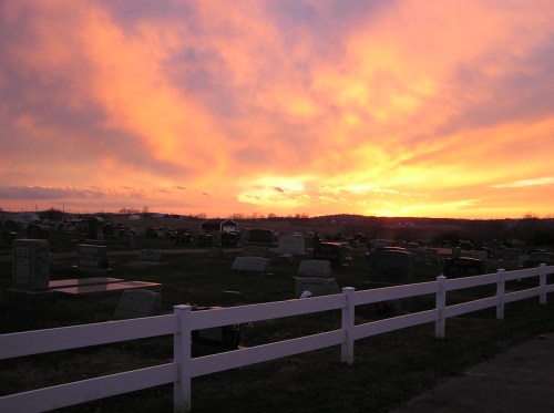A serene cemetery at sunset, with colorful clouds and a white fence in the foreground.
