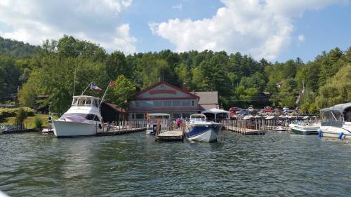A scenic view of a marina with boats docked, surrounded by trees and a building in the background under a blue sky.