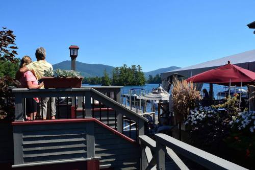 A couple stands on a deck overlooking a lake, surrounded by trees and mountains under a clear blue sky.
