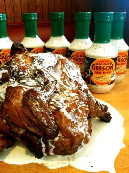 A whole grilled chicken drizzled with white sauce, surrounded by bottles of Big Bob Gibson Bar-B-Q sauce.