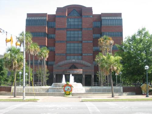 A modern brick government building with a fountain in front, surrounded by palm trees and greenery.