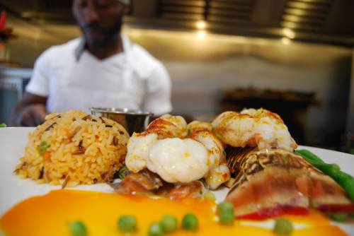 A close-up of a gourmet seafood dish with shrimp, rice, and vegetables, with a chef blurred in the background.
