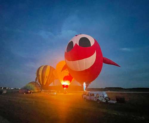 Colorful hot air balloons, including a large red one with a smiling face, illuminated against a twilight sky.
