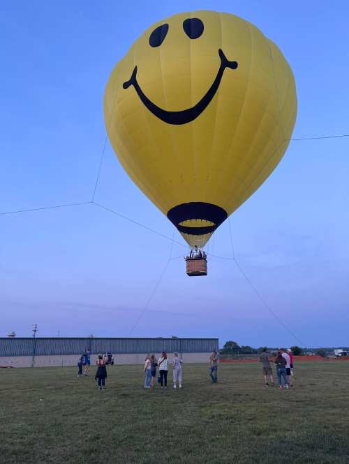A large yellow hot air balloon with a smiley face floats above a grassy field, with people gathered below.