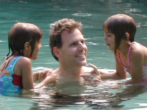 A man smiles while holding two young girls in a swimming pool, enjoying a playful moment together.