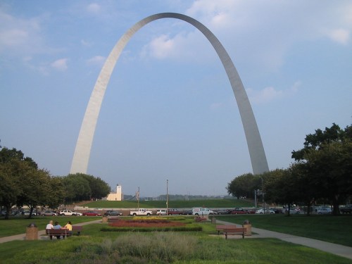 The Gateway Arch towers over a park with benches and trees, under a blue sky with scattered clouds.