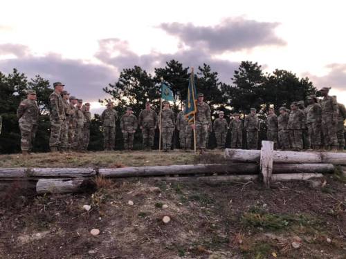 A group of soldiers in military uniforms stands in formation near flags, with trees and a cloudy sky in the background.
