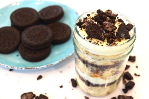 A layered dessert in a jar with cake and cream, topped with crushed cookies, alongside a plate of whole cookies.