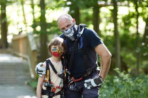A man and a child, both wearing harnesses and masks, smile together in a forested area.