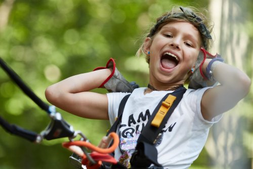 A joyful child in a harness smiles widely, enjoying an outdoor adventure in a green, wooded area.