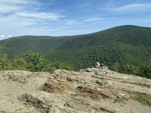 A person relaxes on a rocky ledge, overlooking lush green mountains under a blue sky with scattered clouds.
