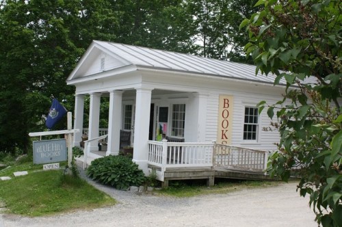 A charming white bookstore with a porch, sign reading "Blue Hill Books," surrounded by greenery and a flag.