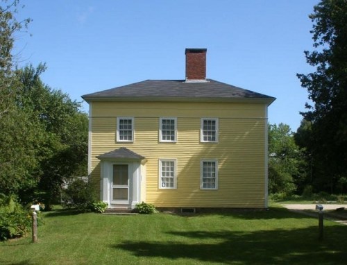 A two-story yellow house with white windows and a front porch, surrounded by green grass and trees under a clear blue sky.