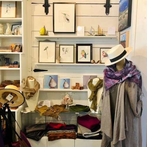 A cozy display of art and clothing, featuring framed bird illustrations, scarves, and hats on mannequins and shelves.