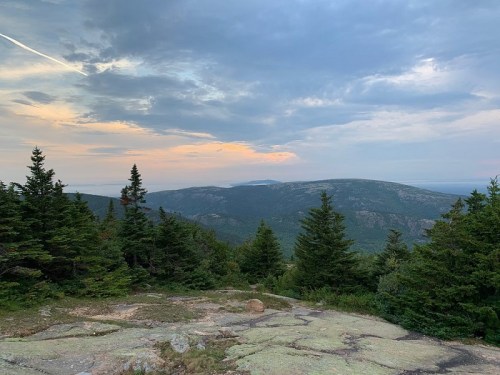 A scenic view of rolling hills and trees under a cloudy sky at sunset, with rocky terrain in the foreground.