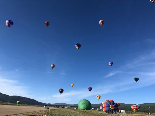 A clear blue sky filled with colorful hot air balloons, with a few on the ground and mountains in the background.