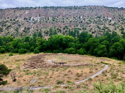 A circular archaeological site surrounded by greenery, with visitors walking along a path in a dry, hilly landscape.