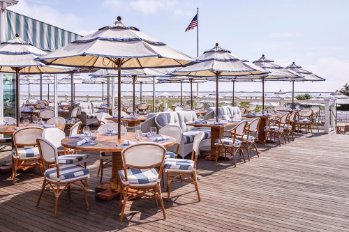 Outdoor dining area with wooden tables, striped umbrellas, and a view of the water under a clear blue sky.