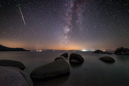 A serene lake at night, with smooth rocks in the foreground and a stunning view of the Milky Way and a shooting star.