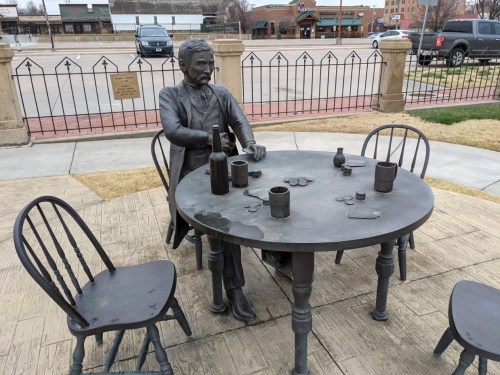 A bronze statue of a man sitting at a table with bottles and poker chips, surrounded by empty chairs.