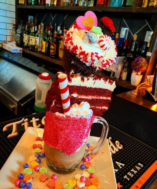 A colorful dessert drink topped with whipped cream, a slice of red velvet cake, and candy decorations on a bar counter.