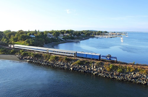 A train travels along a scenic waterfront, with trees and boats in the background under a clear blue sky.