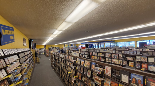 A wide view of a Blockbuster store aisle filled with DVDs and video rentals, with customers browsing.