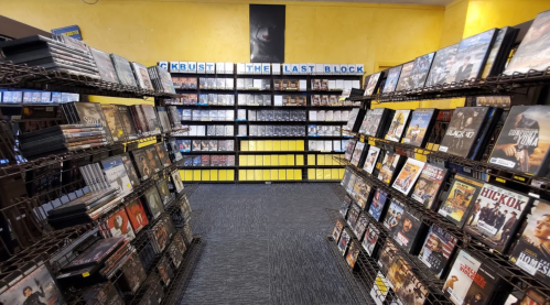A view of a video rental store aisle with shelves filled with DVDs and a yellow wall in the background.