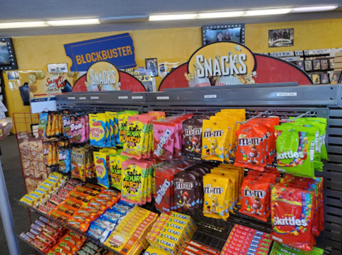 A colorful display of snacks and candy bags in a retro Blockbuster store setting.
